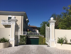 Une entrée fermée par un portail, avec un chemin pavé de pierres menant à un bâtiment doté d’un balcon et d’un pont enjambant un bassin d’eau.