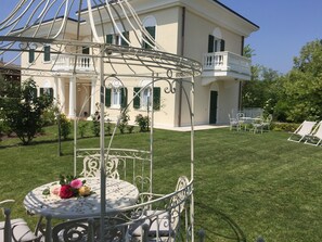 Un belvédère en métal blanc, doté d’une table et de chaises, est entouré d’une pelouse bien entretenue et d’une grande maison avec des balcons.