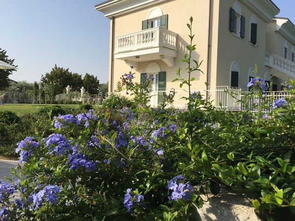 A two-story building with a balcony, surrounded by lush greenery and flowering plants.