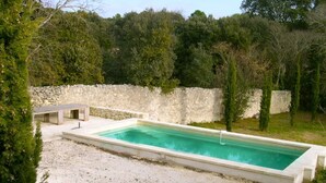 A rectangular swimming pool with a stone wall and a bench in a natural setting.