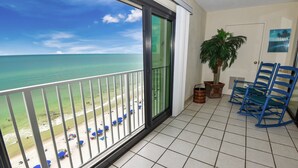 Un balcon donnant sur la plage, un palmier en pot et des fauteuils à bascule bleus.