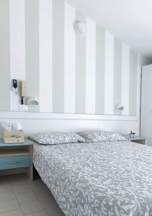 A hotel room with a striped wall, a bed with patterned bedding, a bedside table with a lamp, and a small plant. - Alba B&B Appartamenti (Cattolica)