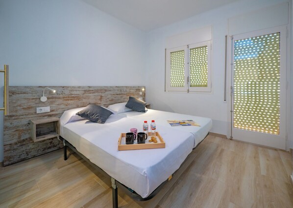 Un dormitorio moderno con cabecera de madera, una mesita de noche con lámpara, una cama con almohadas, una bandeja con alimentos para el desayuno y una ventana con persiana estampada.