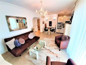 A living room with a brown sofa, two armchairs, a glass coffee table, and a dining area with a white table and chairs.