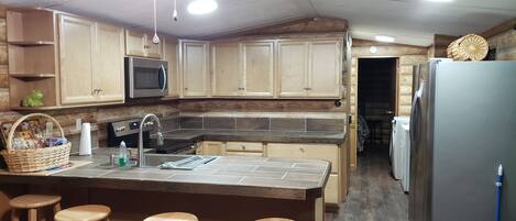 A compact kitchen with wooden cabinets, a microwave, a sink, and a refrigerator. There are three wooden stools at the kitchen island.