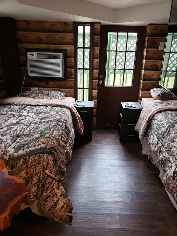 Una habitación tipo cabaña con dos camas, piso de madera, ventana con diseño de celosía y un equipo de aire acondicionado en la pared.