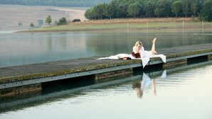 Une personne allongée sur une jetée en bois qui s’avance sur un lac paisible, avec une colline boisée en arrière-plan.
