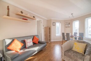 Ein Wohnzimmer mit grauem Sofa, einem Sessel, einem Esstisch mit Stühlen, einem Bücherregal und einem Fenster.