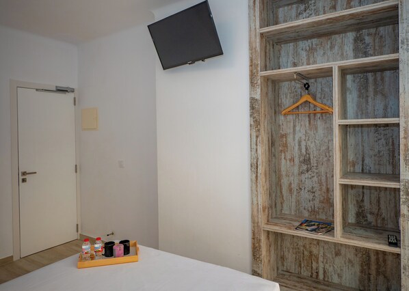 Une pièce avec un lit blanc, une étagère en bois sur laquelle repose un magazine, un porte-vêtements suspendu et une porte avec un interrupteur.