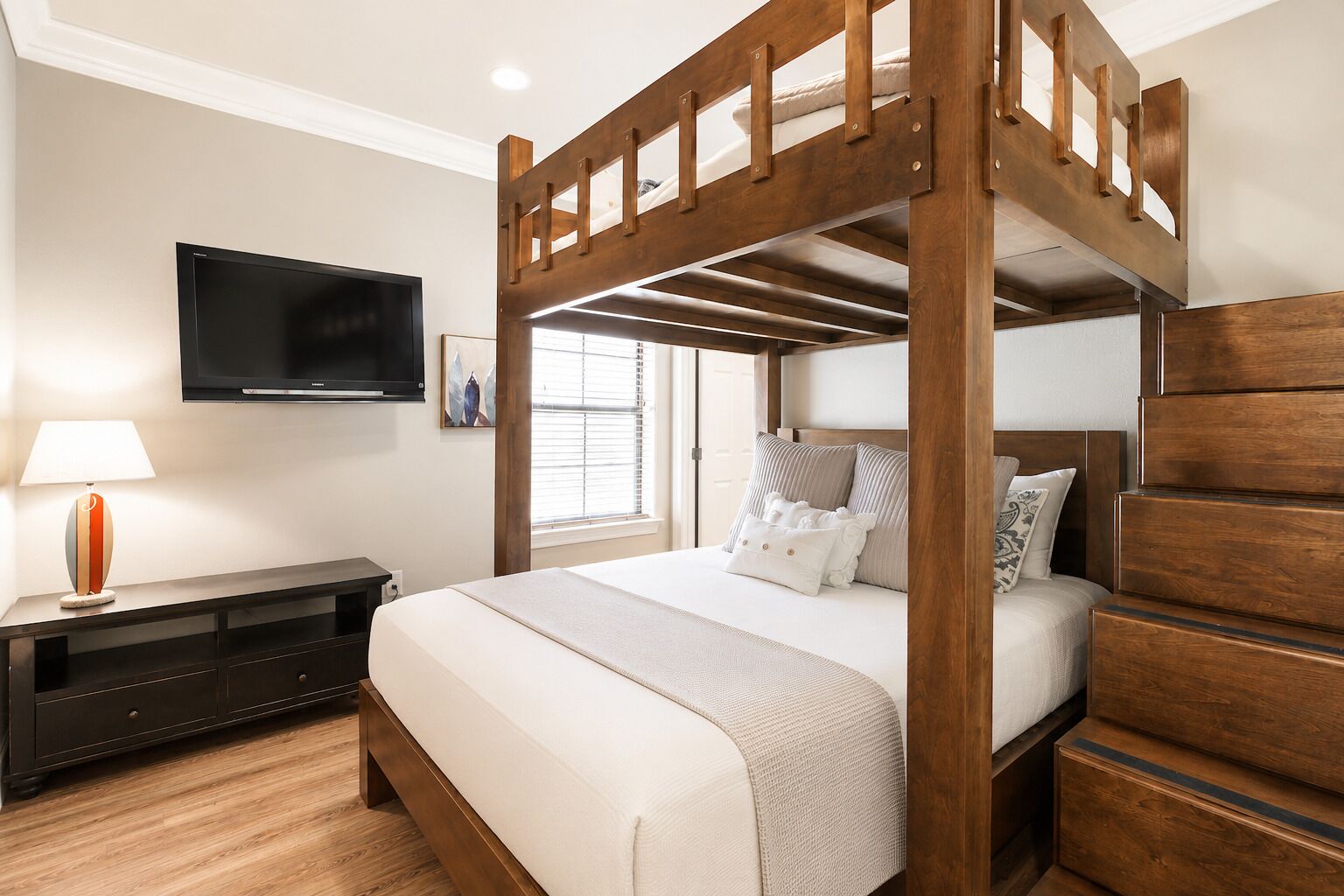 A bedroom with a wooden bunk bed, a flat-screen TV, a bedside table, and a window with blinds.