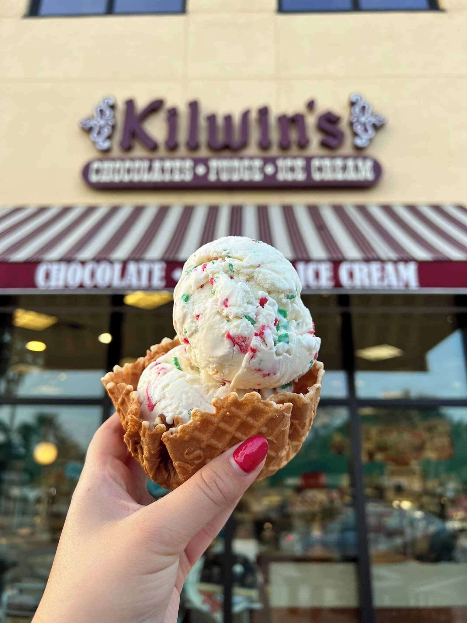 A hand holding a waffle cone with ice cream in front of a storefront with a sign reading "Kilwin's".