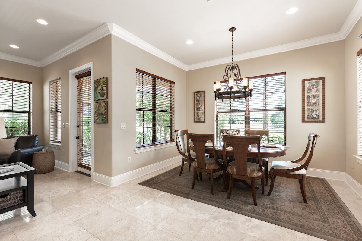 A dining area with a round table and chairs, beige walls, and large windows with blinds.