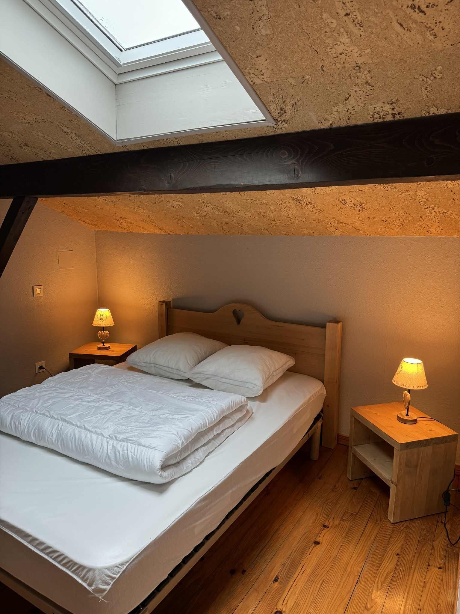 Un dormitorio con tragaluces, vigas de madera, una cama con ropa de cama blanca, dos mesitas de noche con lámparas y una pequeña mesita de noche de madera.