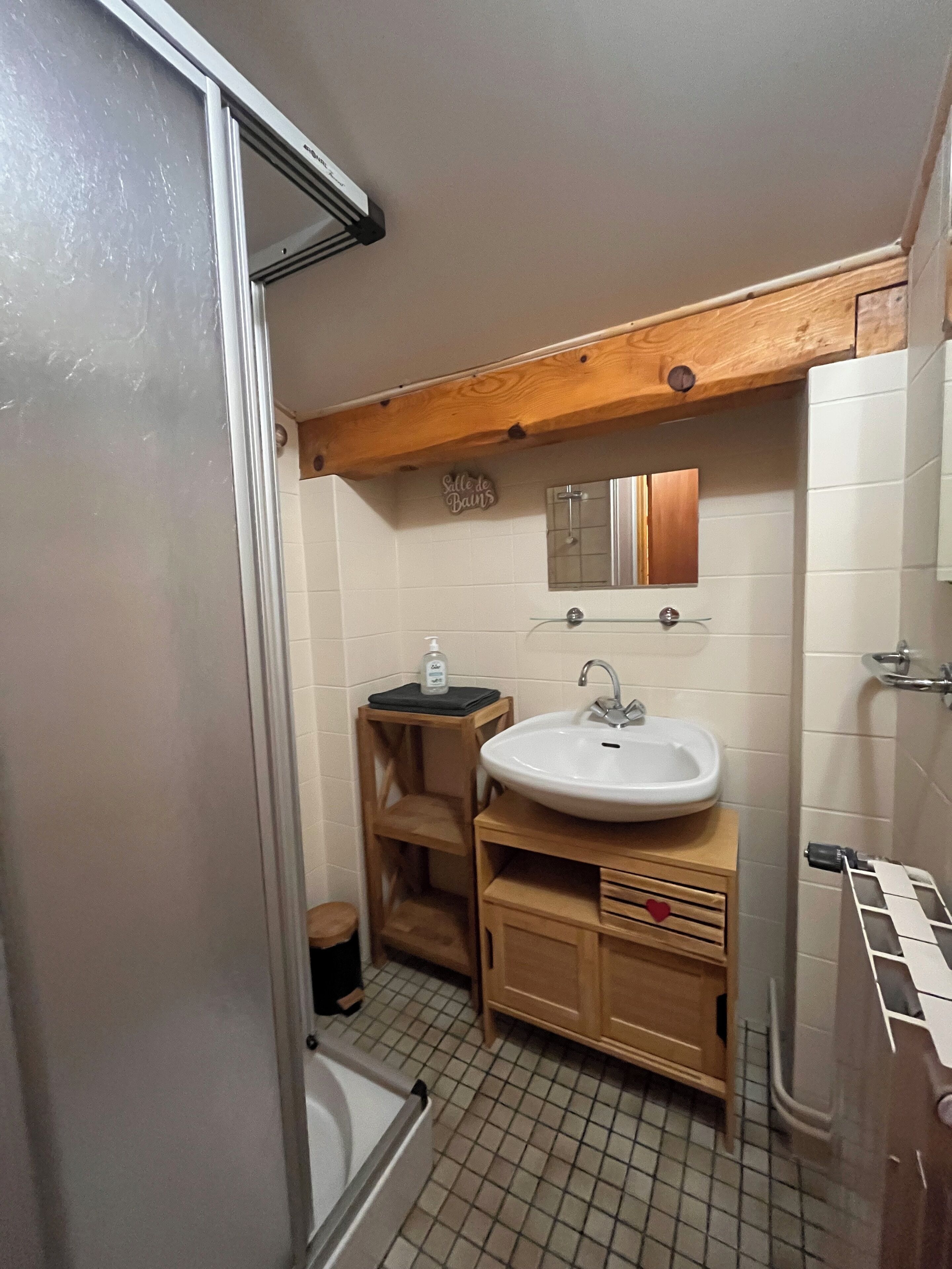 A bathroom with a white sink, wooden cabinets, a small wooden stool, a glass shower enclosure, and a tiled floor.