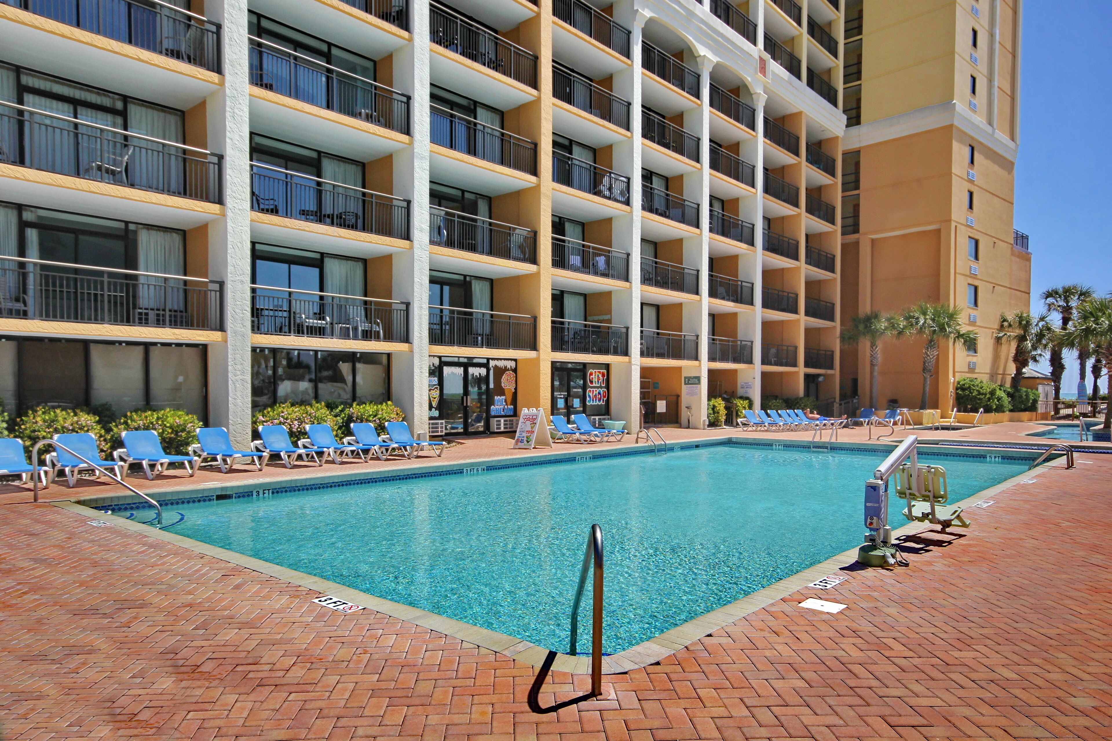 A hotel pool area with a clear pool, blue lounge chairs, and a multi-story building with balconies.