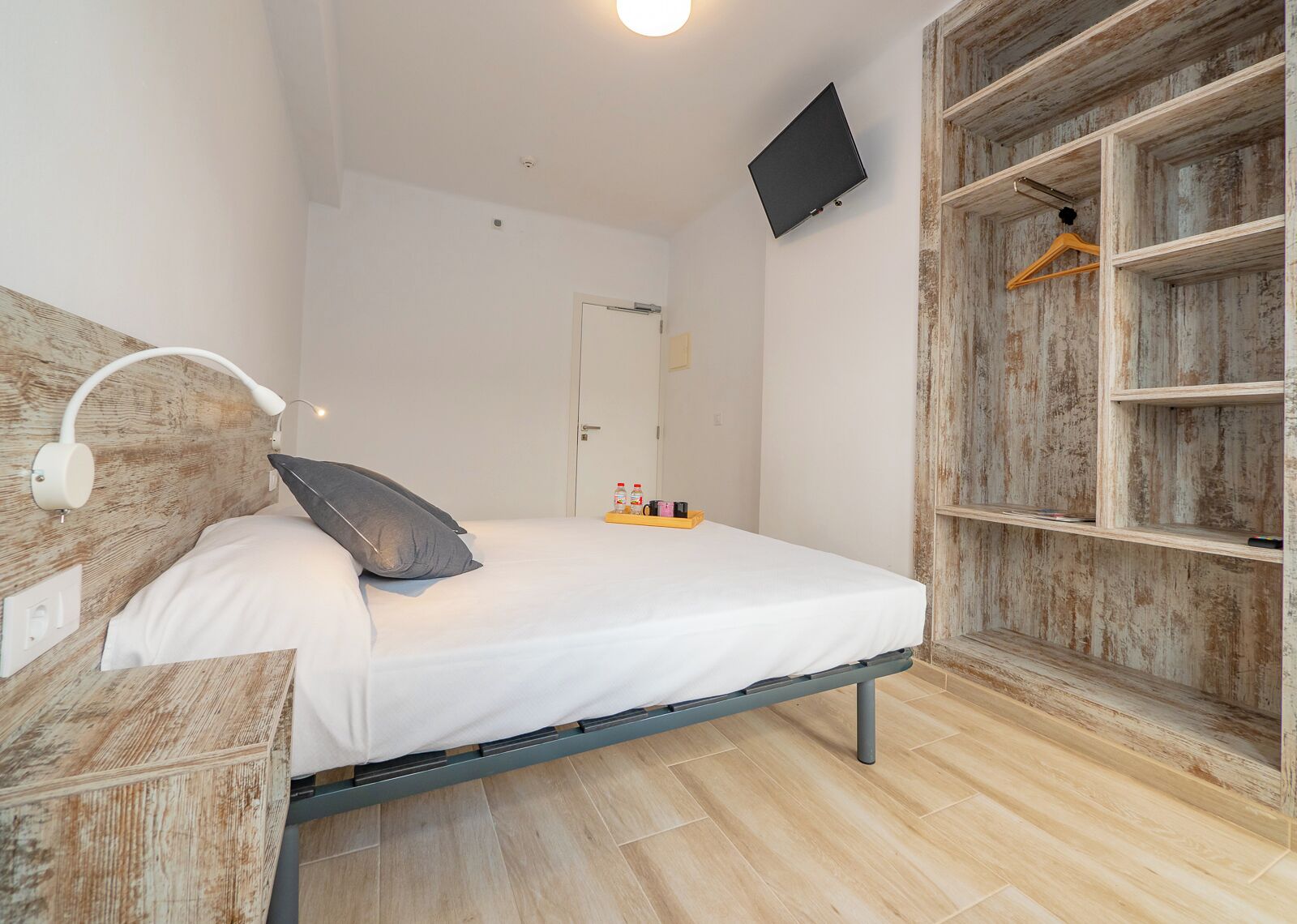 Un dormitorio con cama, mesita de noche, cabecera, televisor montado en la pared y un armario de madera con barra para colgar.