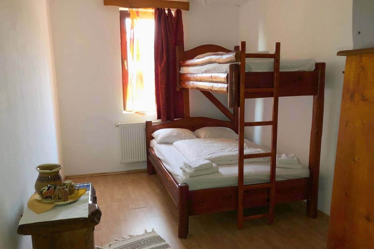 A small, cozy room with a bunk bed, a radiator, a window with curtains, a small table with a vase, and a wooden dresser.