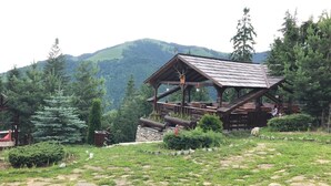 A rustic wooden cabin with a sloped roof, surrounded by lush greenery and mountains in the background.