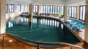 A large indoor swimming pool with clear views of the outdoors through large windows, surrounded by lounge chairs.