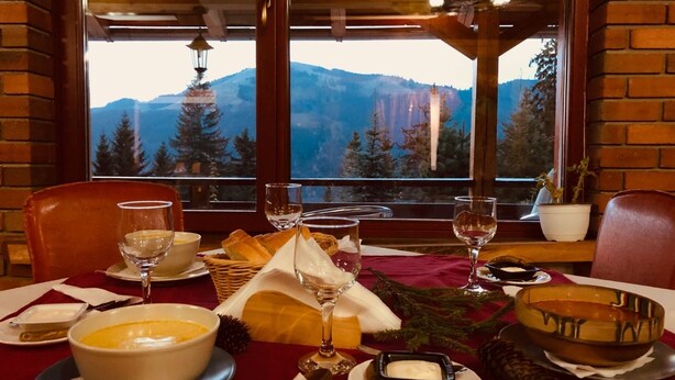 A dining table set with a bowl of soup, bread, wine glasses, and a candle holder, with a mountain view through the window.