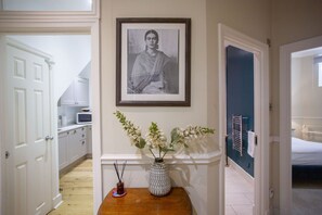 A hallway with a framed photograph on the wall, a wooden table with a vase of flowers, and a bed in the adjacent room. - Quiet, City Garden Apt. Pets Allowed (PL) (Bath)