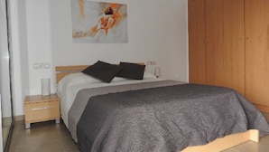 A bedroom with a bed, a wooden wardrobe, a nightstand, a lamp, and a painting on the wall. - Residencial complex les marines  (Roses)