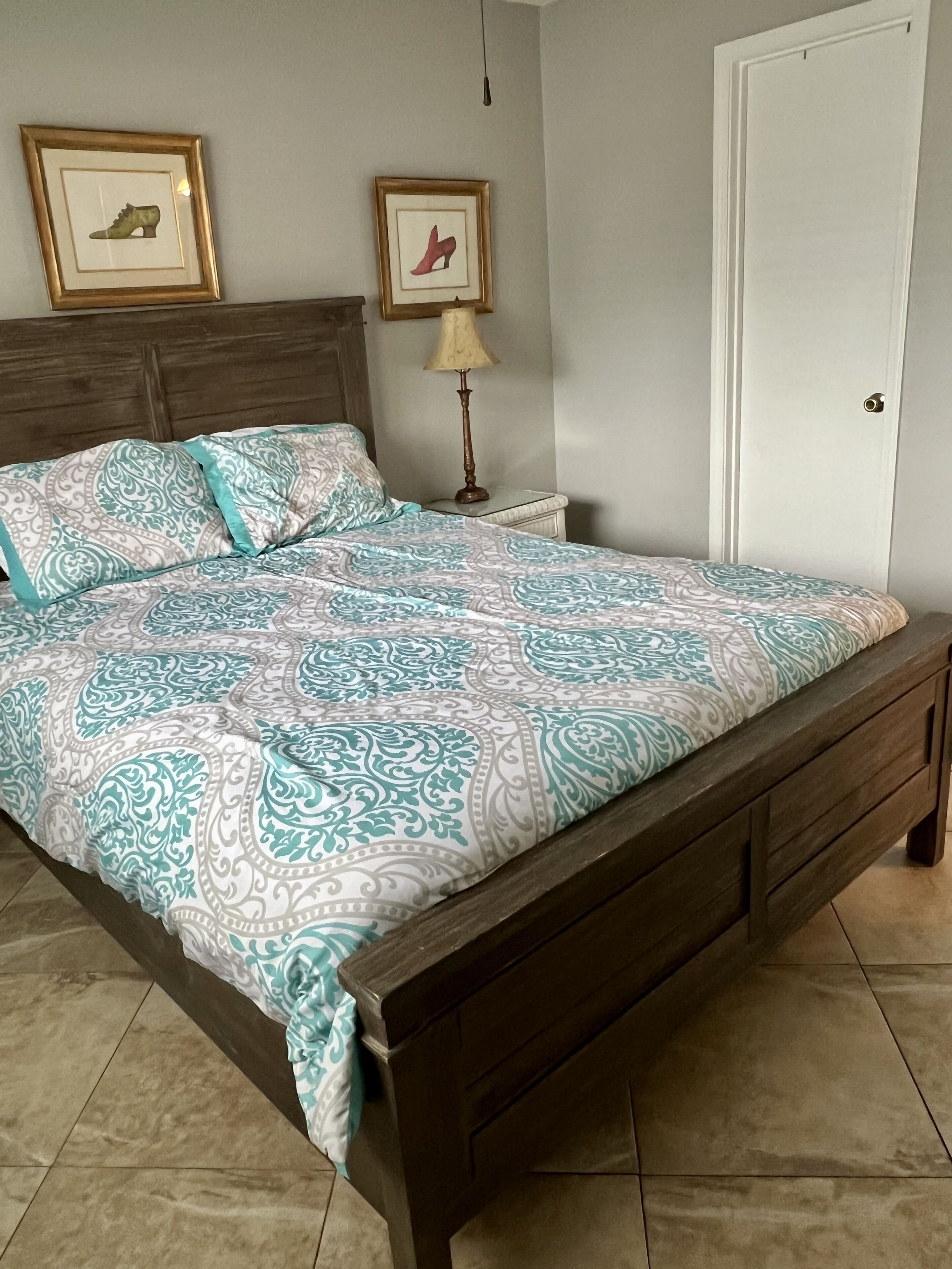 A bedroom with a wooden bed, patterned bedding, a nightstand with a lamp, and two framed pictures on the wall.