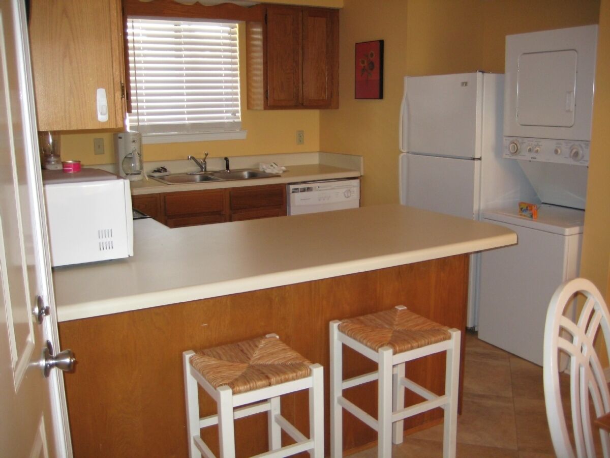 A compact kitchen with wooden cabinets, a white countertop, a sink, a refrigerator, and a washing machine. There are two stools at the counter.