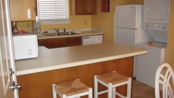 A compact kitchen with wooden cabinets, a white countertop, a sink, a refrigerator, and a washing machine. There are two stools at the counter.