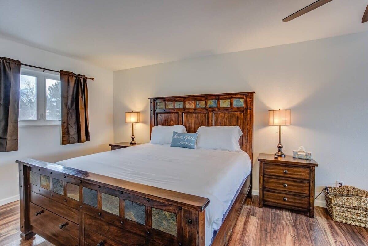 Un dormitorio con una cama de madera, dos mesitas de noche con lámparas, un pequeño aparador y una ventana con cortinas.
