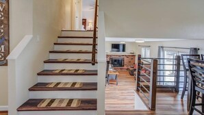Una escalera de madera con barandilla, que conduce a un espacio de estar con chimenea, televisor y mesa de comedor.