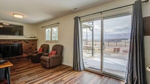 Un salón con chimenea, dos sillones, una mesa y un amplio ventanal corredero que da a un patio exterior.