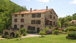 A stone building with a red-tiled roof, multiple windows with shutters, and a small bridge leading to the entrance. - cottage with a view of the river (Le Tech)