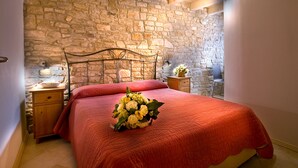 Quarto com parede de pedra, teto de madeira, estrutura de cama de metal, colcha vermelha, mesa de cabeceira com abajur e criado-mudo com um vaso de flores.