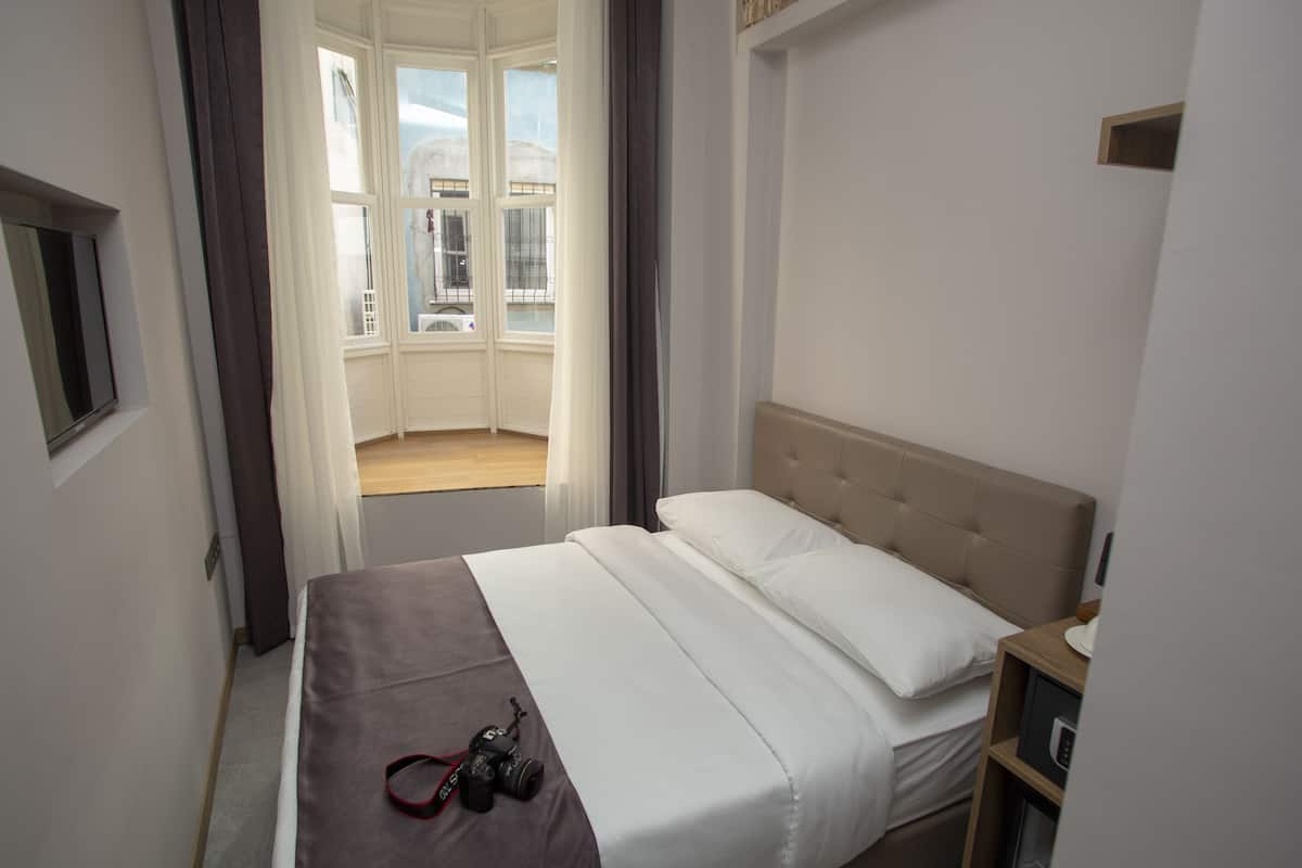 A hotel room with a bed, a camera on the bed, a window with curtains, and a view of buildings outside.