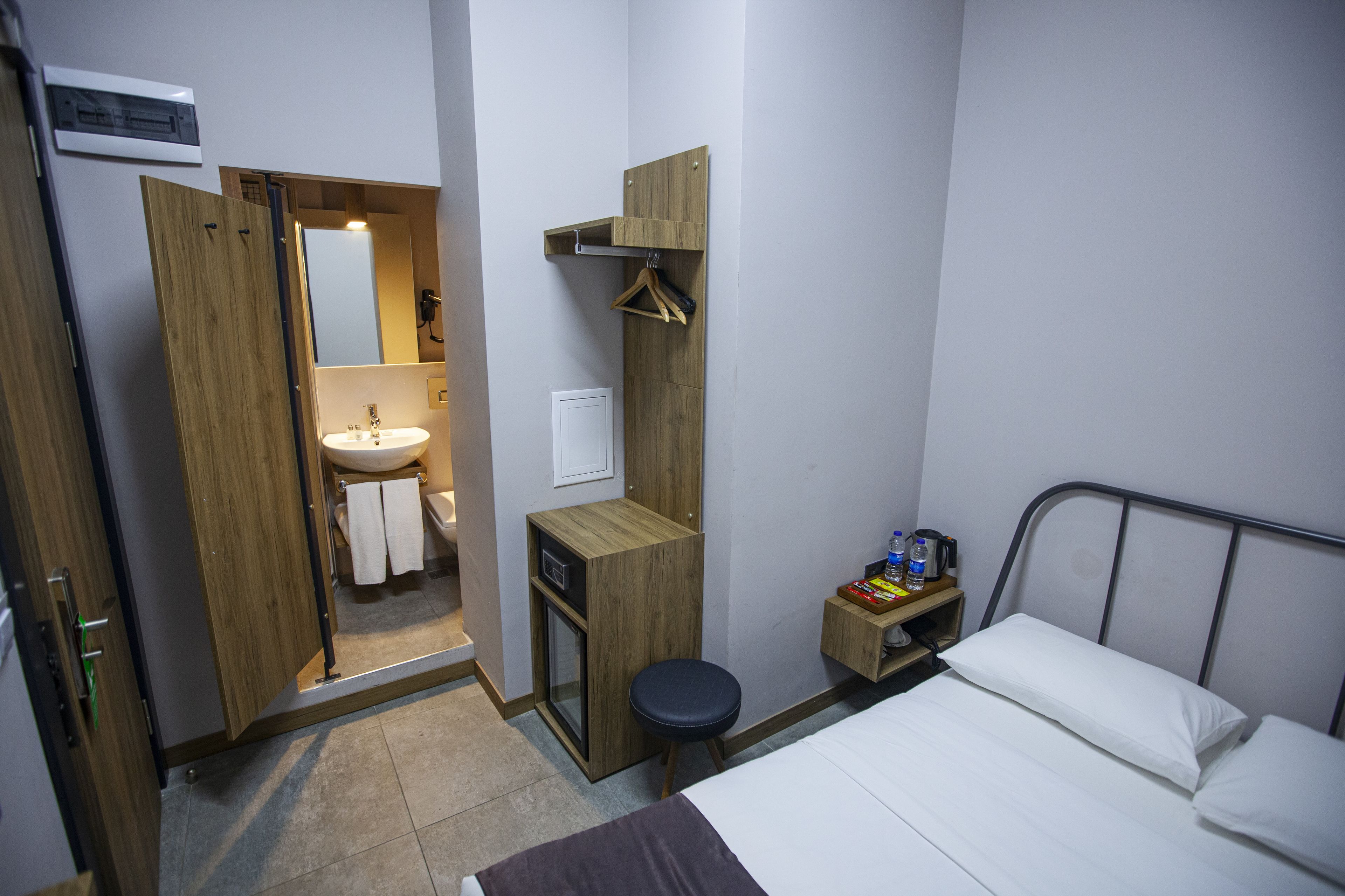Ett kompakt hotellrum med en garderob i trä, ett litet handfat, ett nattduksbord med en vattenkokare och en säng för en person.