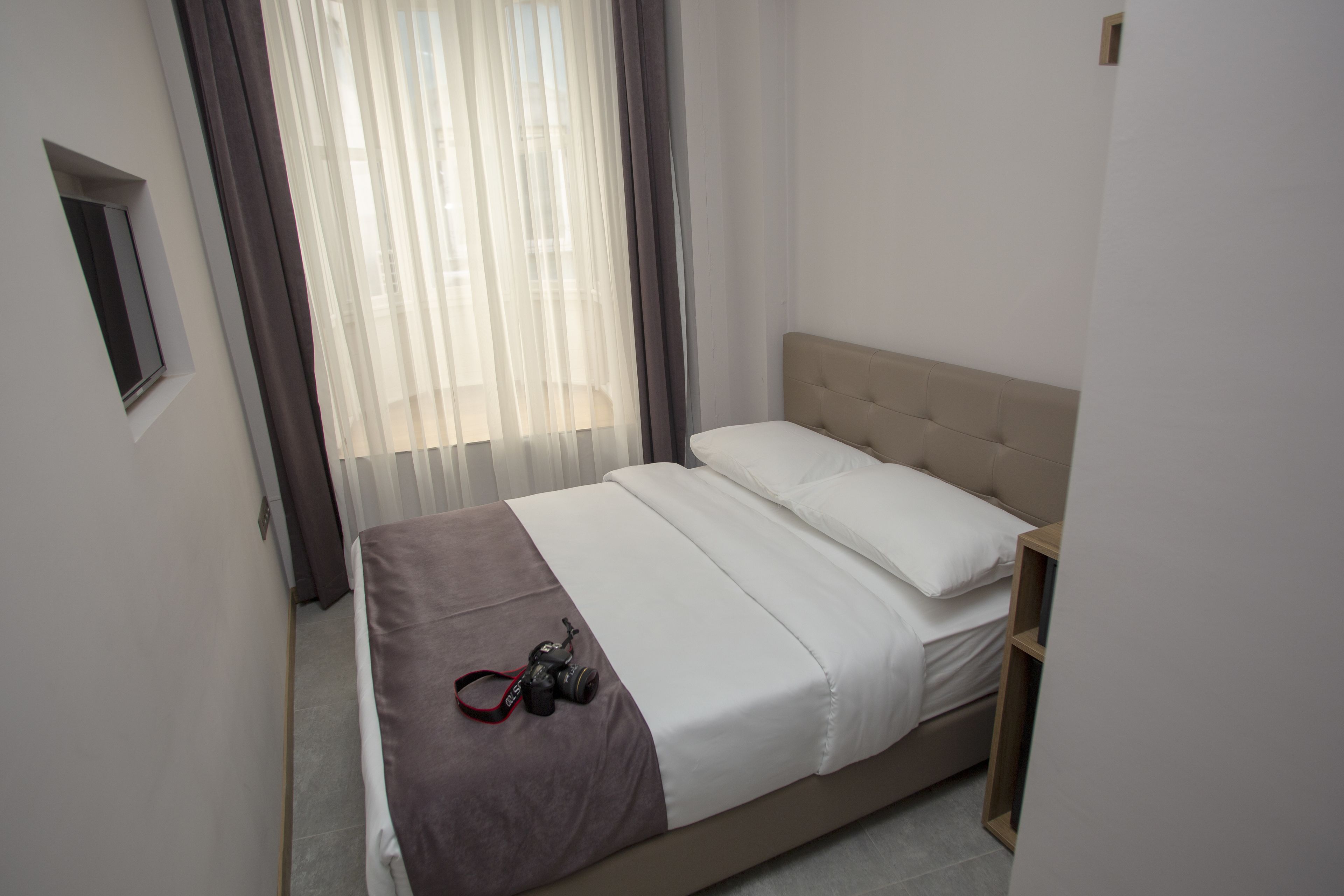 A neatly made bed with a camera on the side, a bedside table, and a window with sheer curtains.