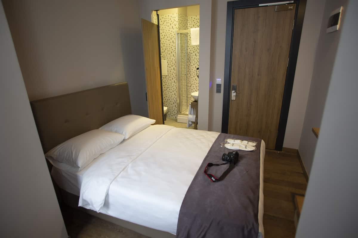 A neatly made bed with white linens and a gray throw blanket, a camera on the bed, and a wooden door in the background.