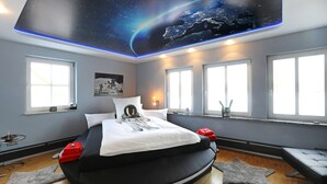 Un dormitorio con una cama, un techo con motivos espaciales, ventanas amplias y una taza con temática de Star Wars en el alféizar.