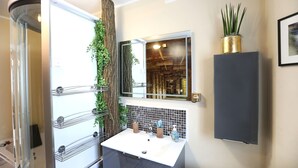 Un baño con cabina de ducha de vidrio, un lavabo blanco con salpicadero de azulejos, un espejo y una planta en maceta.
