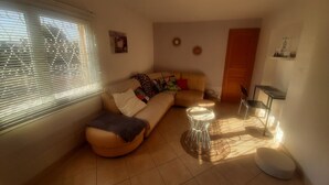 Un salon avec un canapé, un pouf et une petite table. Il y a une fenêtre avec des stores, une porte et un mur orné d’un cadre.