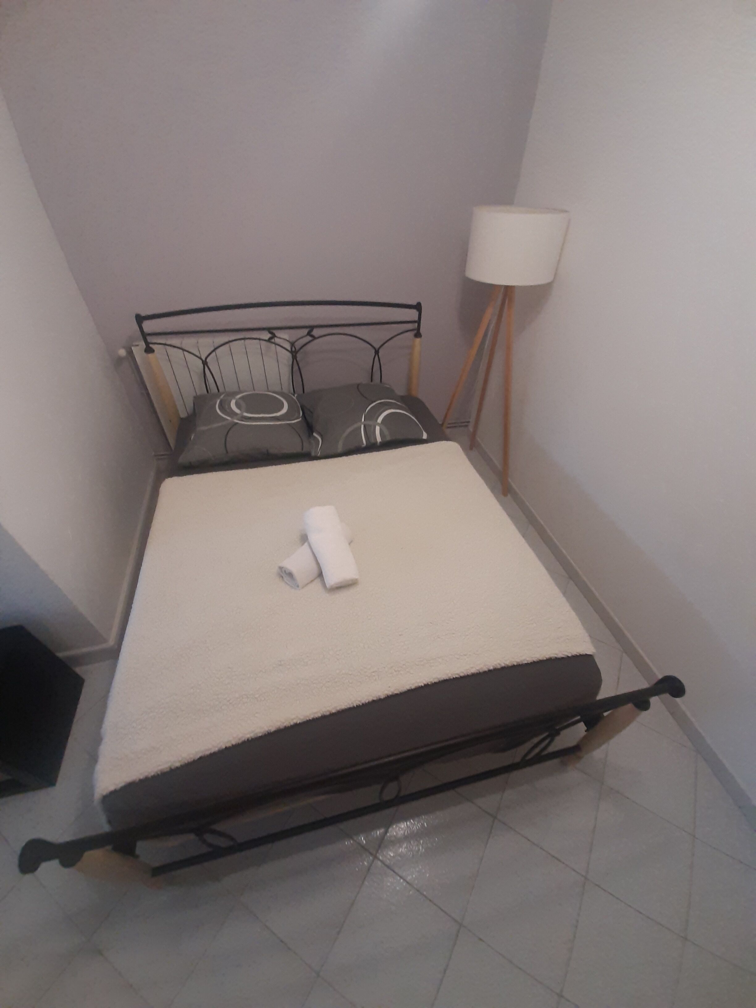 A small, neatly made bed with a white mattress and grey pillows, a white blanket, and a rolled-up towel.