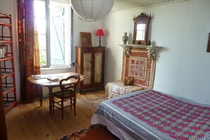 Una stanza con una scrivania in legno, una sedia, un letto con copriletto decorato, un camino con mensola ornata e una finestra con tende.