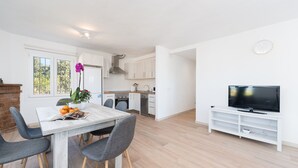 Uma cozinha moderna com armários brancos, uma mesa de jantar com cadeiras, uma TV sobre um suporte branco e uma janela com vista para árvores.
