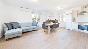 Uma sala de estar moderna com um sofá cinzento, uma mesa de jantar com cadeiras, e uma área de cozinha com fogão e armários.