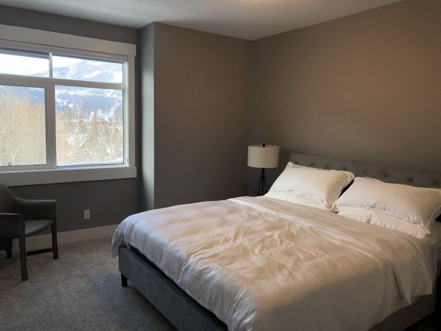 A bedroom with a large bed, a chair, a nightstand with a lamp, and a window with a view of mountains.