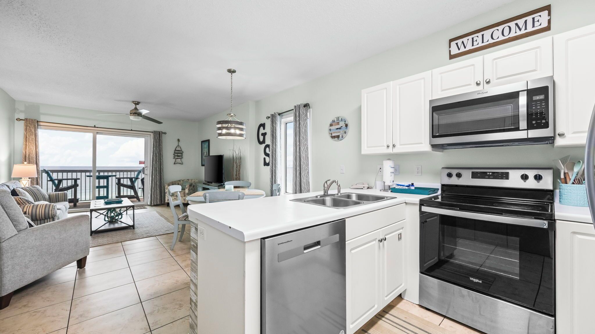 A modern kitchen with white cabinetry, a built-in microwave, and an oven. There is a dining area with a table and chairs, a sofa, and a balcony with a view.