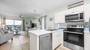 Una cocina moderna con armarios blancos, microondas integrado y horno. Hay un comedor con mesa y sillas, un sofá, y un balcón con vistas.