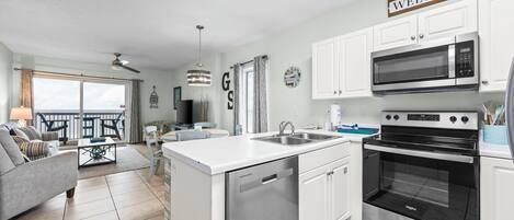 A modern kitchen with white cabinetry, a built-in microwave, and an oven. There is a dining area with a table and chairs, a sofa, and a balcony with a view.