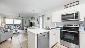 Una cocina moderna con armarios blancos, microondas integrado y horno. Hay un comedor con mesa y sillas, un sofá, y un balcón con vistas.